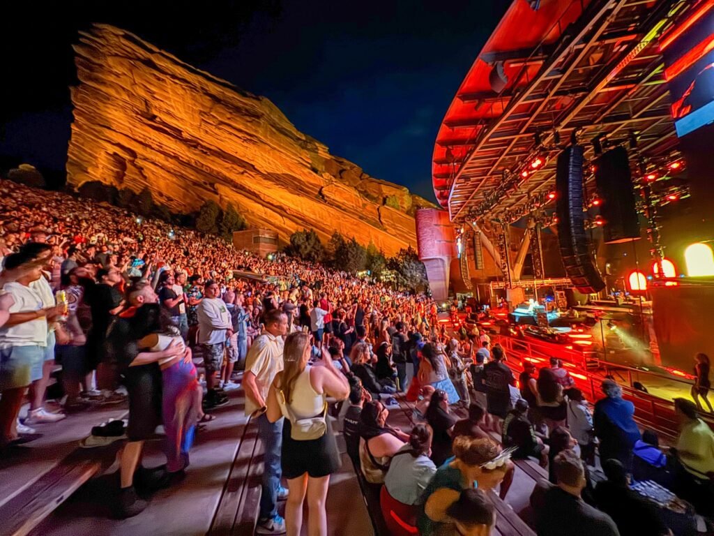 Crowded audience at Red Rocks Amphitheatre during a live concert under colorful stage lights. A canyon wall illuminated in orange glows beside the energetic crowd.