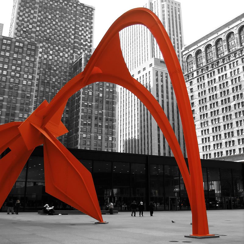 A striking red abstract sculpture stands out against a monochrome cityscape. The bold colors highlight the contrast between art and architecture.