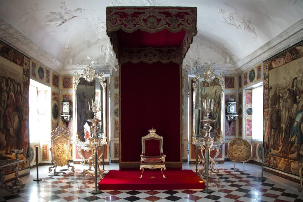 A grand room featuring a red throne under an ornate canopy, surrounded by richly decorated walls and a checkered marble floor.