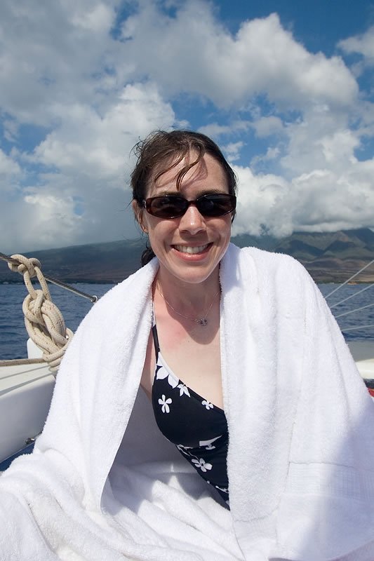 A woman wrapped in a white towel, wearing sunglasses and a floral swimsuit, smiles on a boat with a scenic backdrop of mountains and a partly cloudy sky.