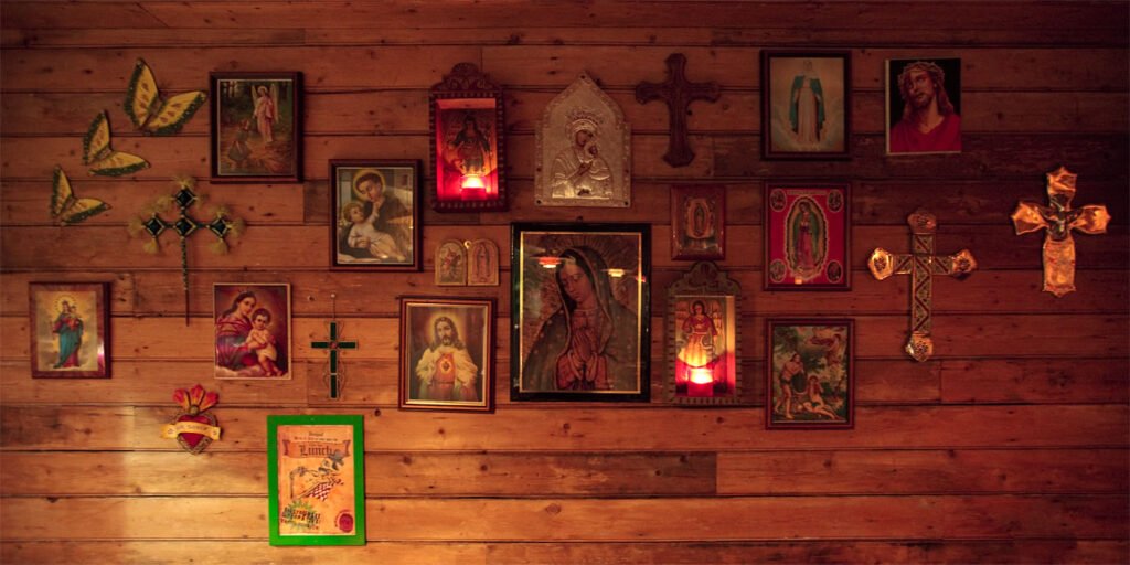 A warm-toned wooden wall adorned with framed religious icons, crosses, and lit candles creating a serene spiritual atmosphere.