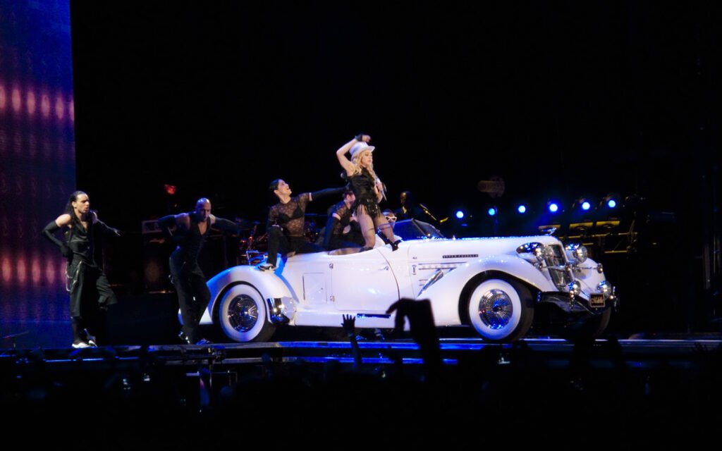 A vintage white car with dancers performing around it under blue stage lights.