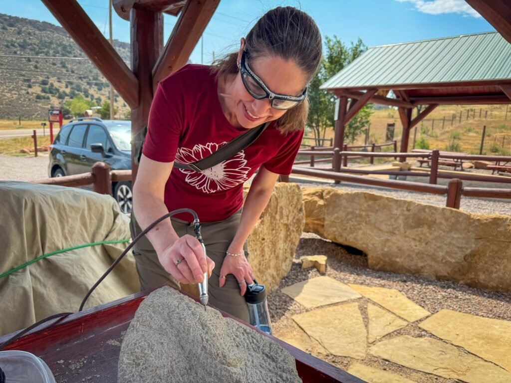 A person carving a rock under a shaded structure with mountains in the background.