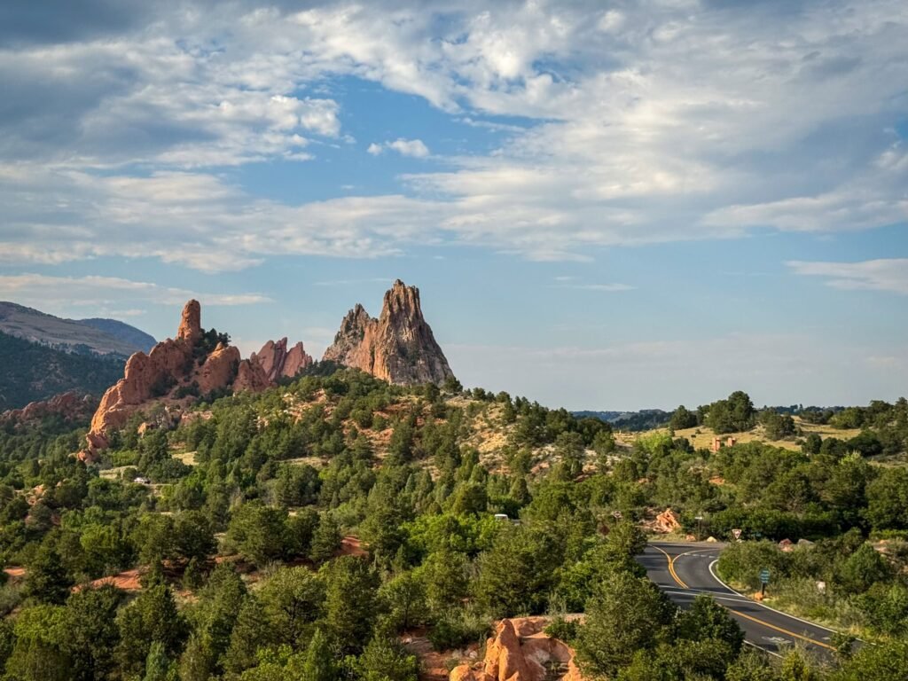 A scenic view featuring prominent red rock formations rising above a lush forested area. A winding road cuts through the greenery under a partly cloudy sky.