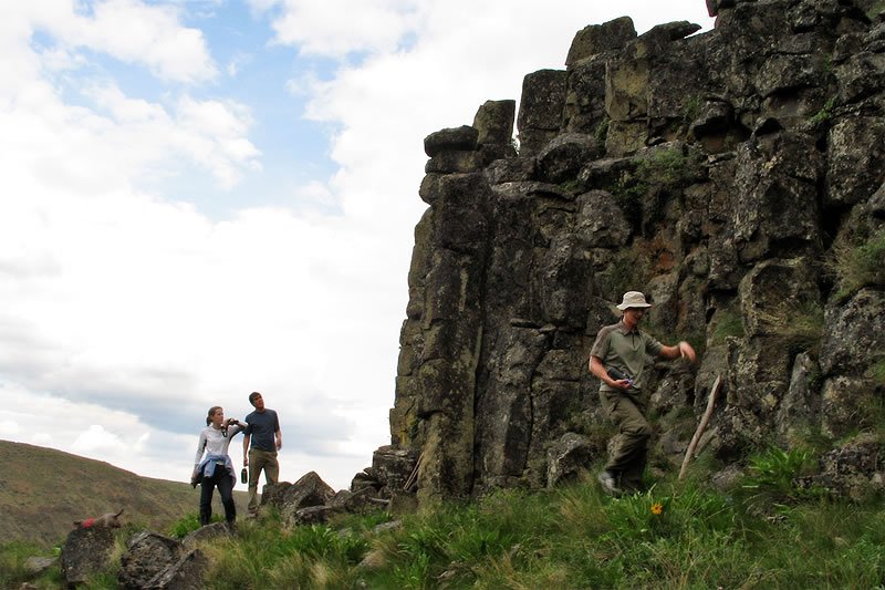 A couple and another person are hiking near large, moss-covered rock formations in a lush green landscape under a partly cloudy sky.