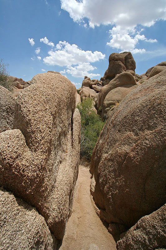 A narrow pathway flanked by giant boulders under a blue sky with scattered clouds.