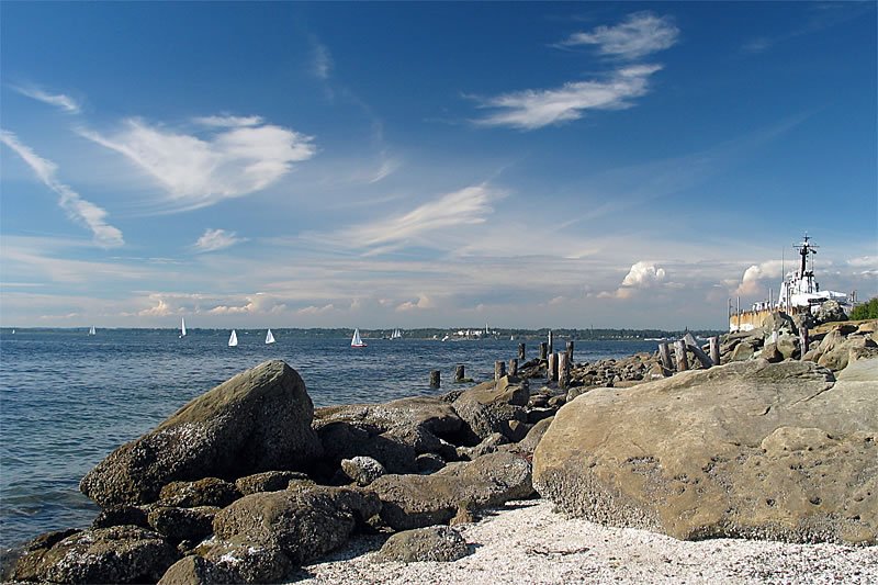 A scenic view of a rocky shoreline meeting the calm blue water, with several sailboats visible in the distance under a partly cloudy sky.