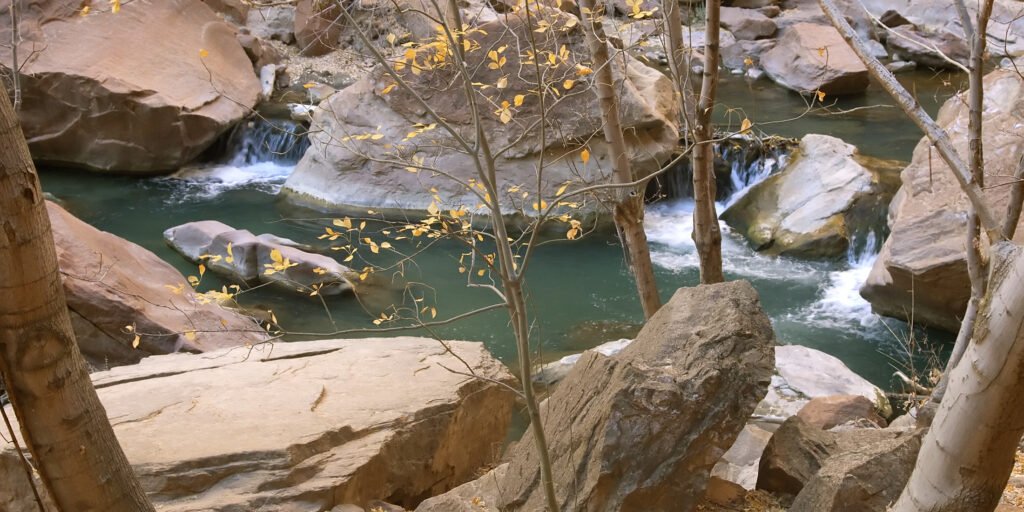 A serene stream flows gently among large boulders and autumn-colored trees.