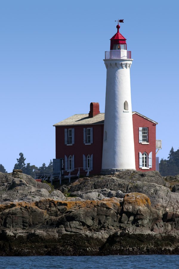 A historic red brick lighthouse with a white tower and red roof stands on a rocky island surrounded by calm blue waters under a clear sky.