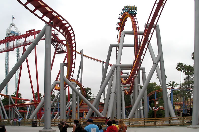 A vibrant roller coaster with red tracks and gray supports twists through an amusement park, offering exhilarating loops and steep drops for adventurous visitors.