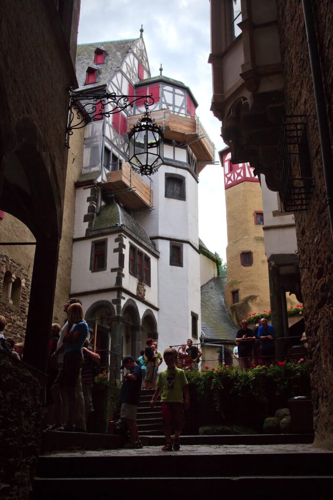 Crowds walking through a narrow alleyway in a European-style village with white and yellow buildings and red shutters.