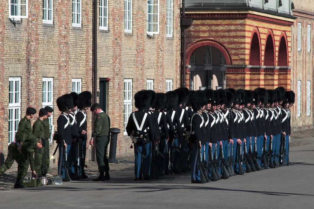 A line of royal guards in traditional black uniforms and tall bearskin hats stand at attention. The scene features a historic brick building with elegant arched windows in the background.