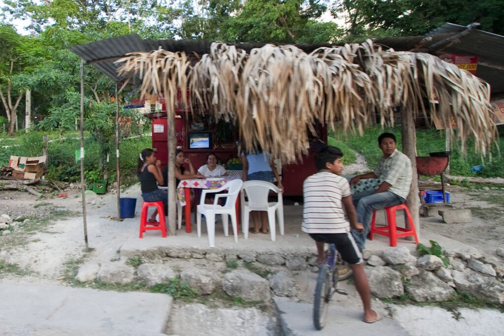 A rustic thatched shutter stands in a serene outdoor setting, with people sitting on red stools beneath it, surrounded by natural greenery.