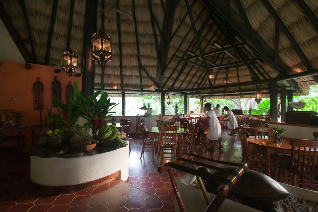 A spacious dining hall featuring a thatched roof and warm lighting. The room is furnished with wooden tables and chairs, surrounded by lush greenery.