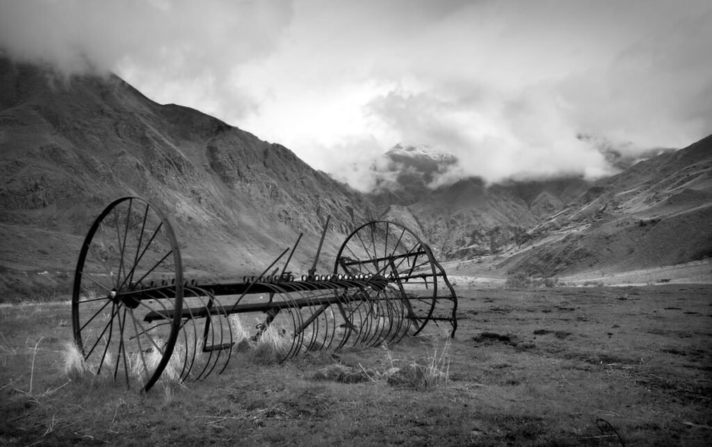 A black and white photograph featuring an old farm plow set against a rugged mountainous backdrop under a cloudy sky.