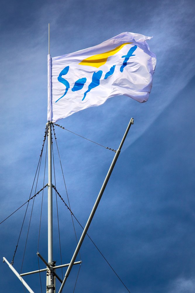 A white flag with blue and yellow details flies atop a sailboat mast against a clear blue sky.