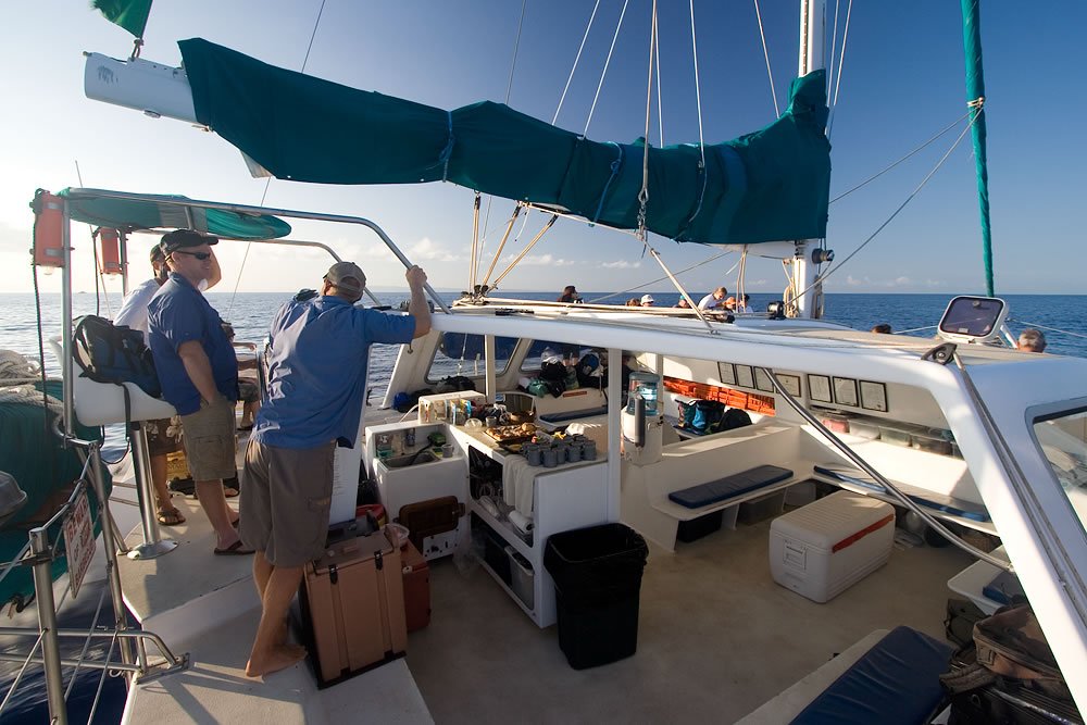 A couple enjoys a sunny day out at sea, navigating a catamaran with a well-equipped deck area.