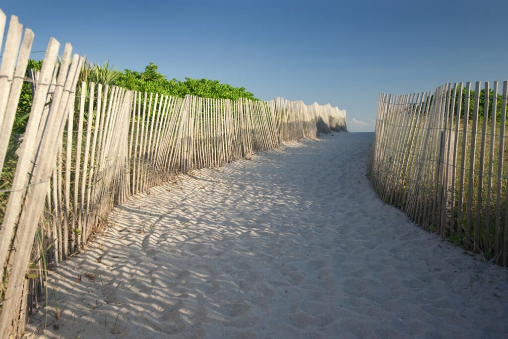 A sandy beach path shaded by wooden fences, leading towards the horizon under a clear blue sky. The fences create dappled shadows on the sand.
