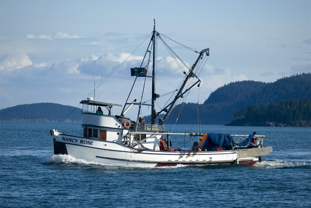 A fishing boat named 'Saint Athan' sails through blue Pacific Northwest waters, with forested hills in the background under a partly cloudy sky.