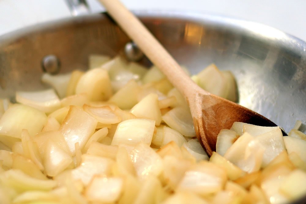 A stainless steel pan with chopped onions being stirred by a wooden spoon.