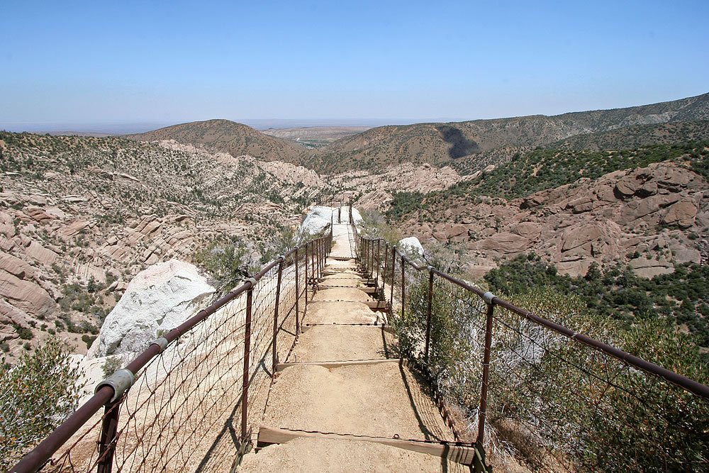 A scenic staircase path with metal railings winding through a rugged hillside. The view overlooks expansive valleys and distant mountains under a clear sky.