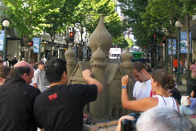 People gather around a large outdoor sculpture on a tree-lined street. The scene is vibrant with activity and greenery.