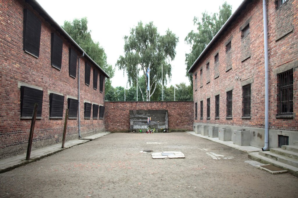 A historic brick courtyard with barred windows and gravel floor, surrounded by two-story buildings under a cloudy sky. Rustic charm captured through weathered textures and muted colors.