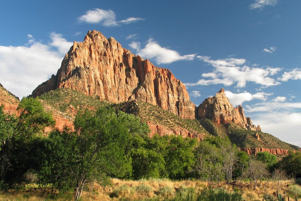 Bright red rock peaks rise dramatically against a blue sky. Green trees and bushes dot the base of the rugged mountain slopes.