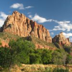 Bright red rock peaks rise dramatically against a blue sky. Green trees and bushes dot the base of the rugged mountain slopes.