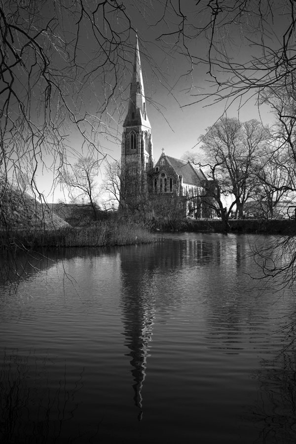 A black and white photograph of a serene cemetery with a prominent steeple reflected in the still water.