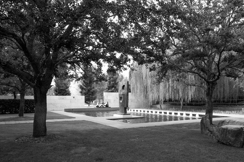 A tranquil pool surrounded by trees and a statue, captured in black and white.