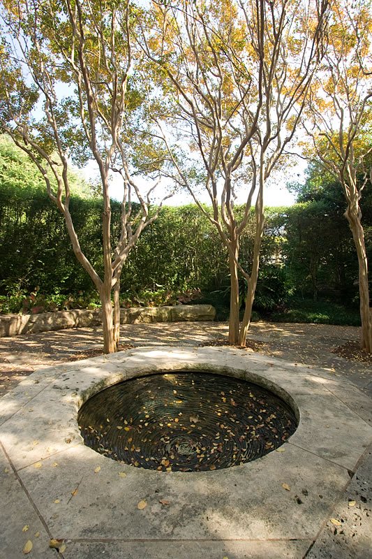 A tranquil water fountain surrounded by trees and foliage, with reflections of the surrounding greenery on its surface.