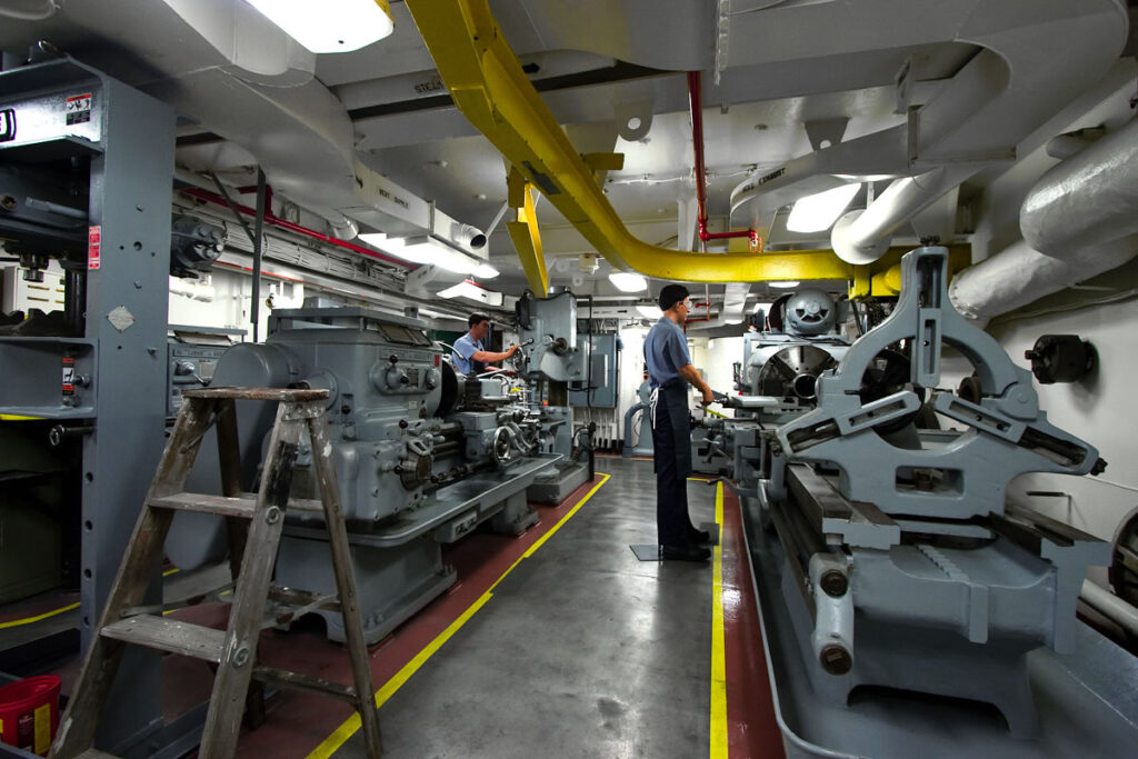 A detailed view of a ship's engine room featuring large machinery, yellow pipes, and crew members. The scene highlights the industrial setting with various equipment and tools visible.