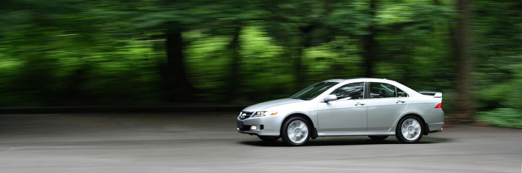 A sleek silver car is captured in motion, driving along a road flanked by lush green trees.