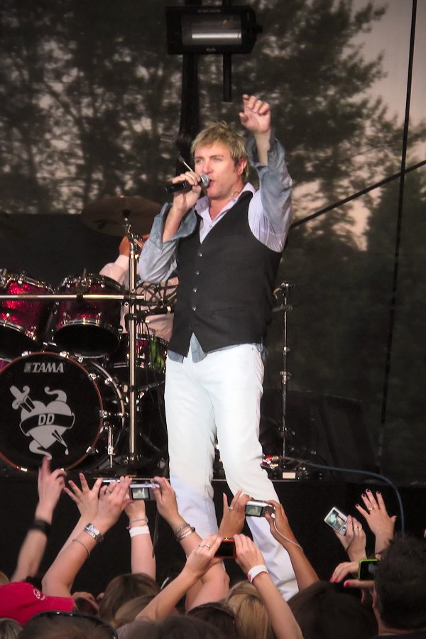 A blond singer in a vest sings into a microphone while raising a hand to the crowd.