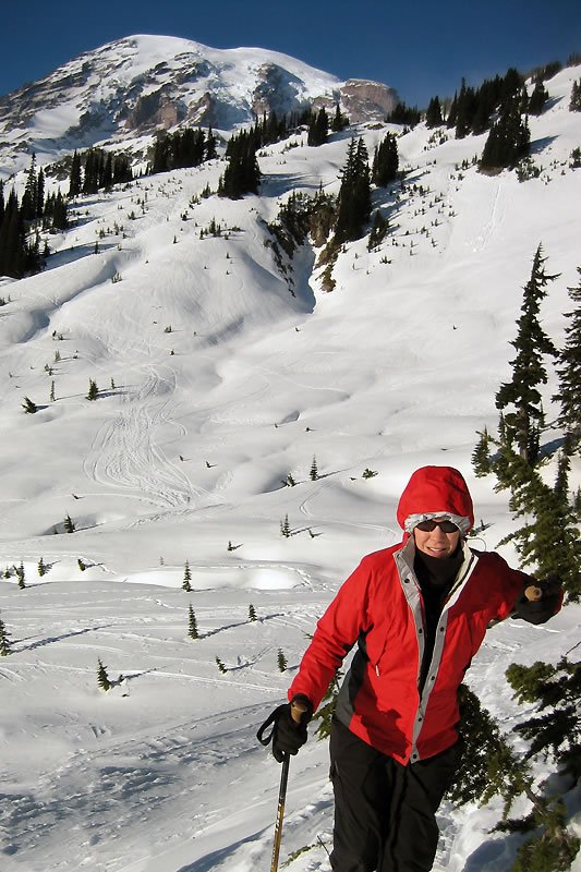 A person dressed in a red jacket and hat stands on a snowy mountain slope with ski poles in hand, surrounded by pine trees and a clear blue sky.