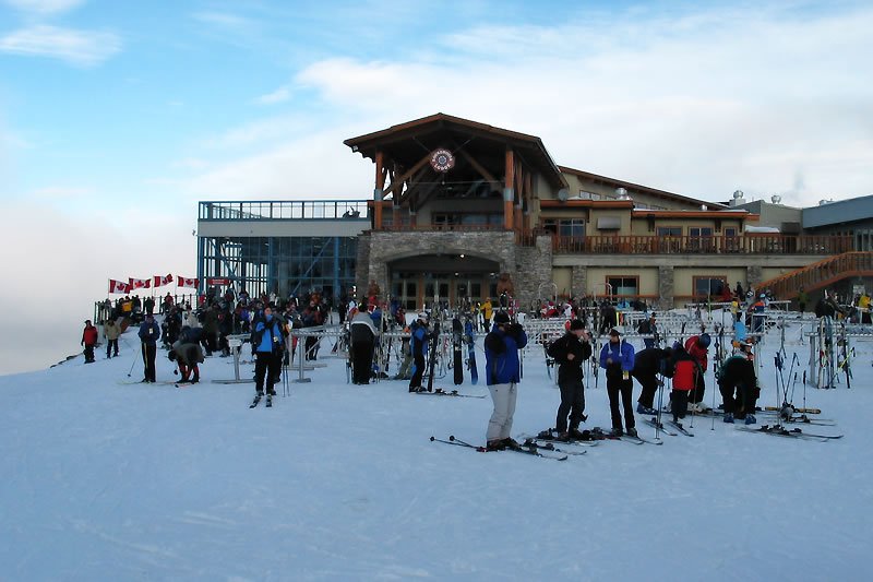A group of skiers gathered near a mountain lodge with a clear blue sky above. Ski equipment is visible on the snowy ground as people prepare for the slopes.
