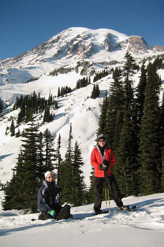 A couple enjoys skiing and snowboarding on a scenic mountain trail surrounded by tall pine trees under a clear blue sky. The majestic peak looms in the background, showcasing a breathtaking winter landscape.