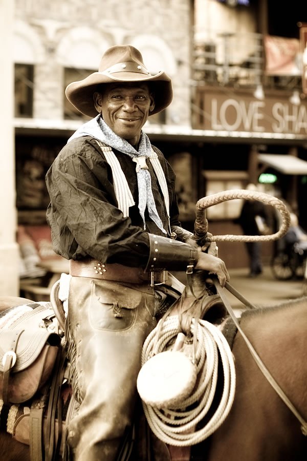 A cowboy in a wide-brimmed hat smiles while sitting on a horse, holding a lasso