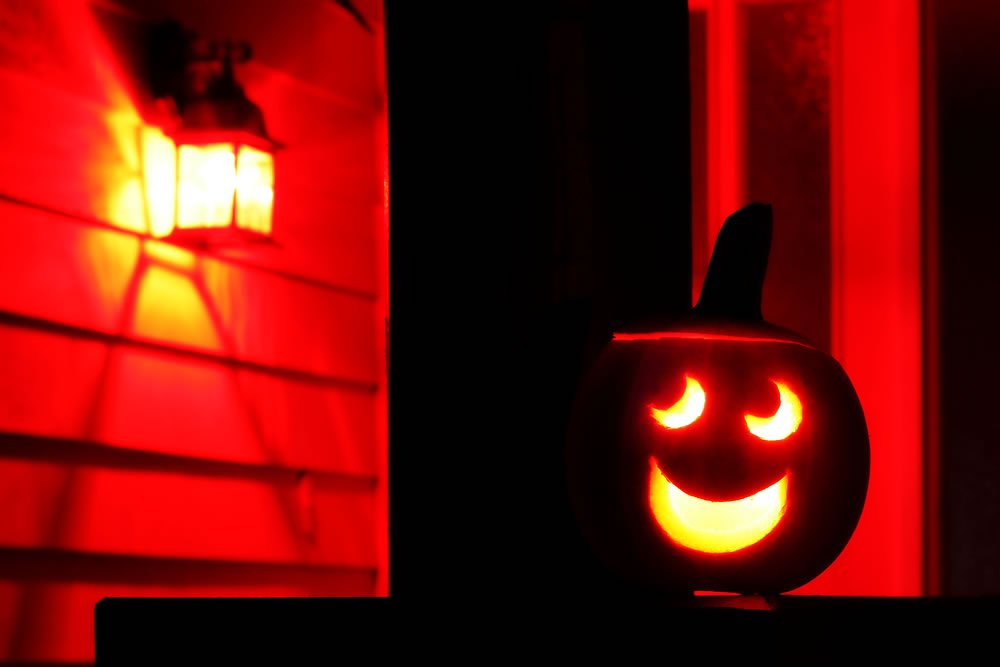 A jack-o'-lantern with a smiling face glows warmly beside a red-lit wall and lantern.