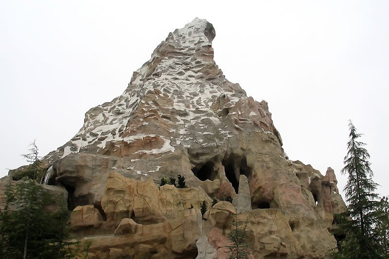 A striking snow-covered rock formation surrounded by lush greenery and tall evergreen trees under an overcast sky.