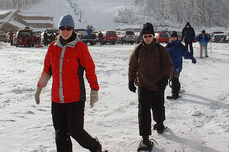 A couple snowshoeing through a snowy landscape with other people and vehicles in the background.
