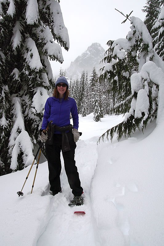 A person snowshoeing through a serene, snow-covered forest trail surrounded by tall evergreen trees.
