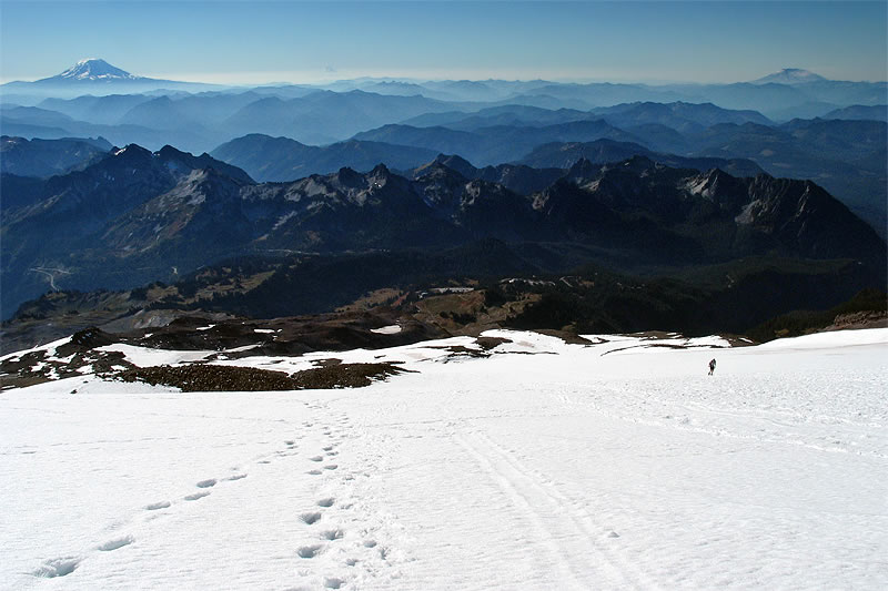 Snow-covered mountain landscape with a trail of footprints leading into the distance. Distant peaks and a clear sky complete the scene.