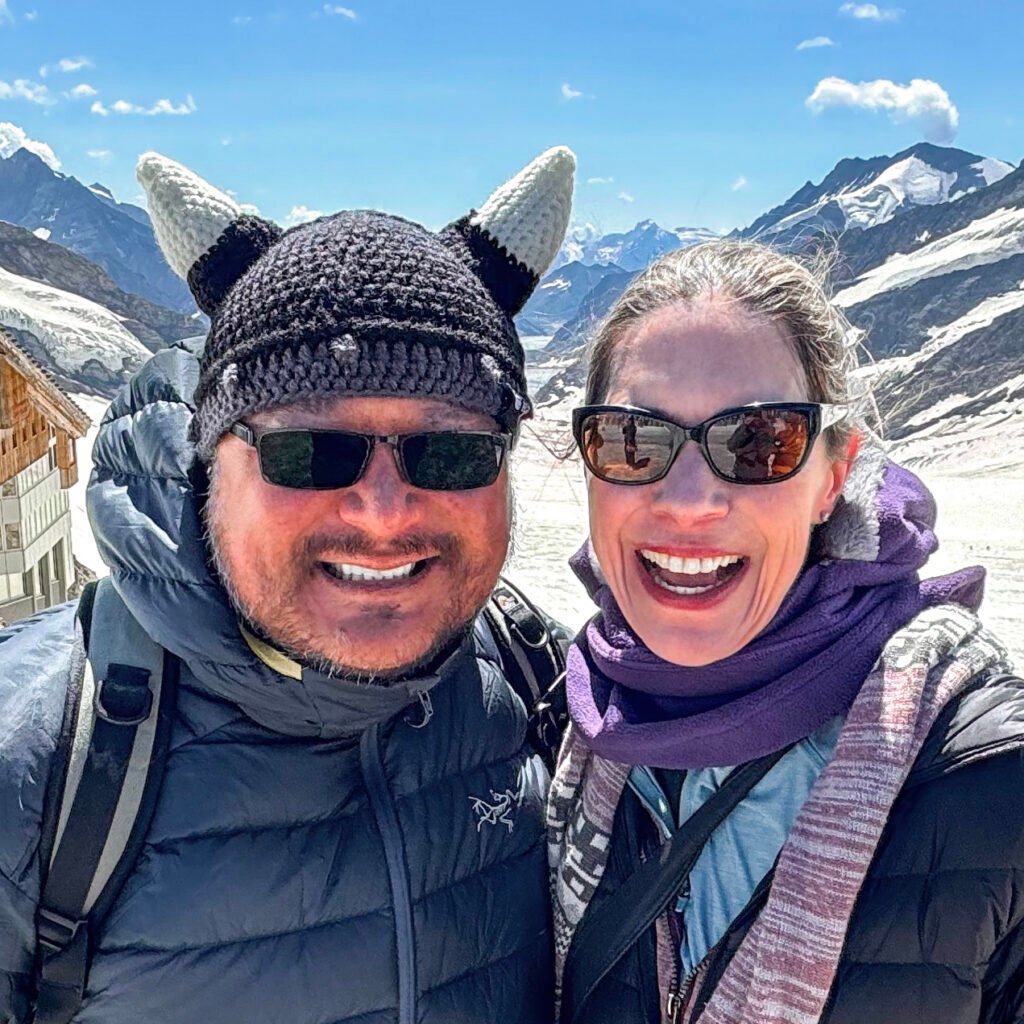 John and Kristie enjoying snowy mountain views wearing winter gear and sunglasses