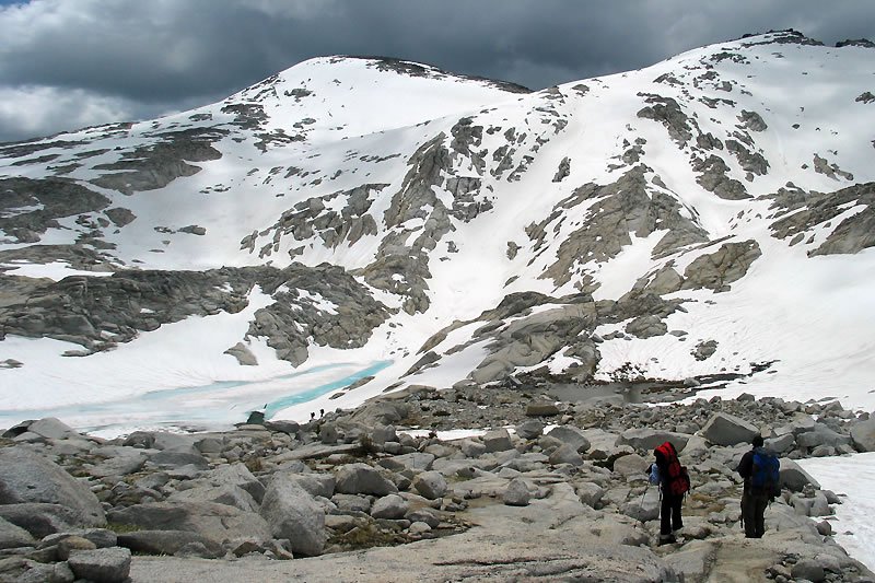 Two hikers traverse a rocky mountain trail under a dramatic cloudy sky.
