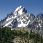 A majestic mountain with snow-covered peaks rises above a forested slope under a clear blue sky.