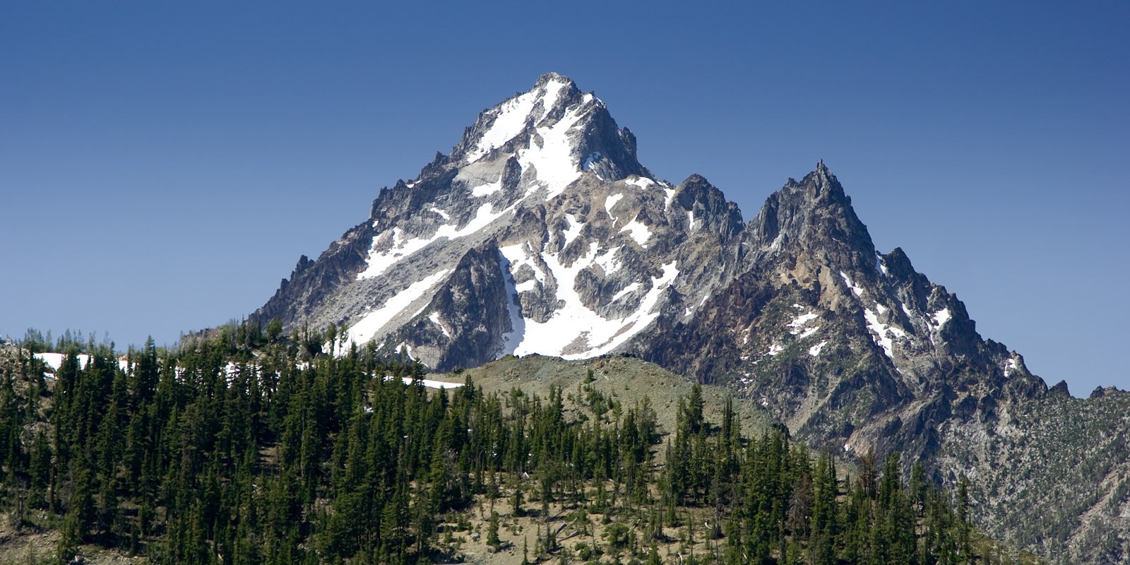 Navaho Peak A majestic mountain with snow-covered peaks rises above a forested slope under a clear blue sky.