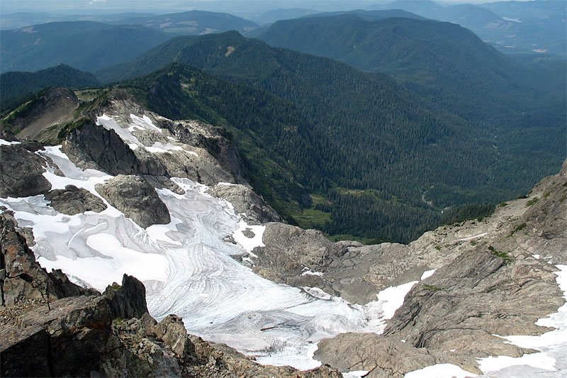 A person stands at the top of a snowy hill, surrounded by tall evergreen trees. The scene captures the serene beauty of the Pacific Northwest winter landscape.