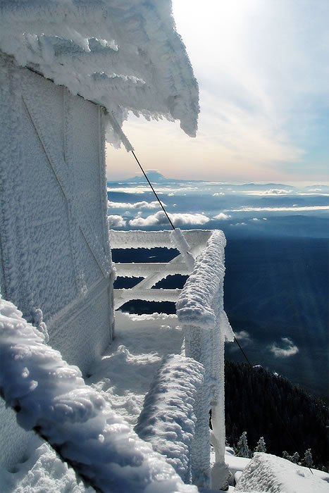 A balcony covered in thick snow with a breathtaking view of the clouds below. The scene captures the serene beauty of a mountain winter wonderland.